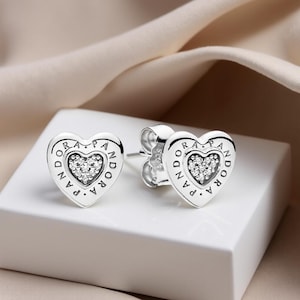 Peut inclure: Une paire de boucles d'oreilles clous en forme de cœur argentées. Chaque boucle d'oreille présente un motif en forme de cœur avec le mot "PANDORA" gravé sur le bord. Le centre de chaque cœur est rempli de petits cristaux transparents. Les boucles d'oreilles sont présentées sur une surface blanche.
