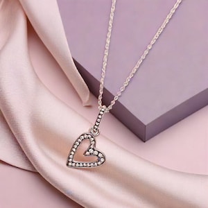 May include: A silver necklace with a heart-shaped pendant. The pendant is encrusted with small, clear crystals around the edges. The necklace chain is a delicate, twisted design. The background features a soft pink fabric and a purple surface.