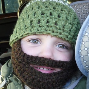 May include: A child wearing a green crocheted hat with a white pom pom and a brown crocheted beard.