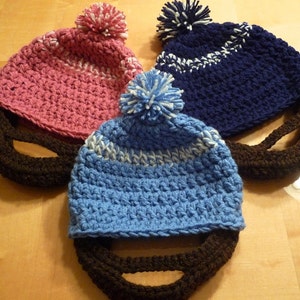 Puede incluir: Tres gorros de ganchillo con barbas de ganchillo marrones. Los gorros son rosa, azul y azul marino con pompones blancos.