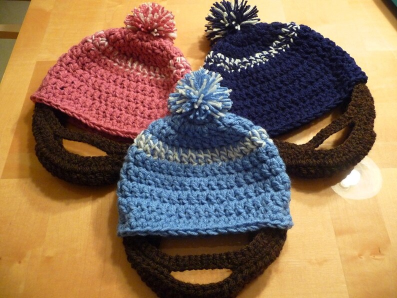 Puede incluir: Tres gorros de ganchillo con barbas de ganchillo marrones. Los gorros son rosa, azul y azul marino con pompones blancos.