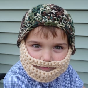 Puede incluir: Un ni&ntilde;o con un gorro de punto de camuflaje y una barba de ganchillo a juego. El gorro y la barba son de color beige y marr&oacute;n.