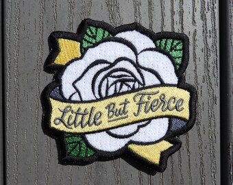 May include: Embroidered patch featuring a white rose with black outlines and green leaves. A yellow banner across the rose reads "Little But Fierce" in black script. The patch has a black border.