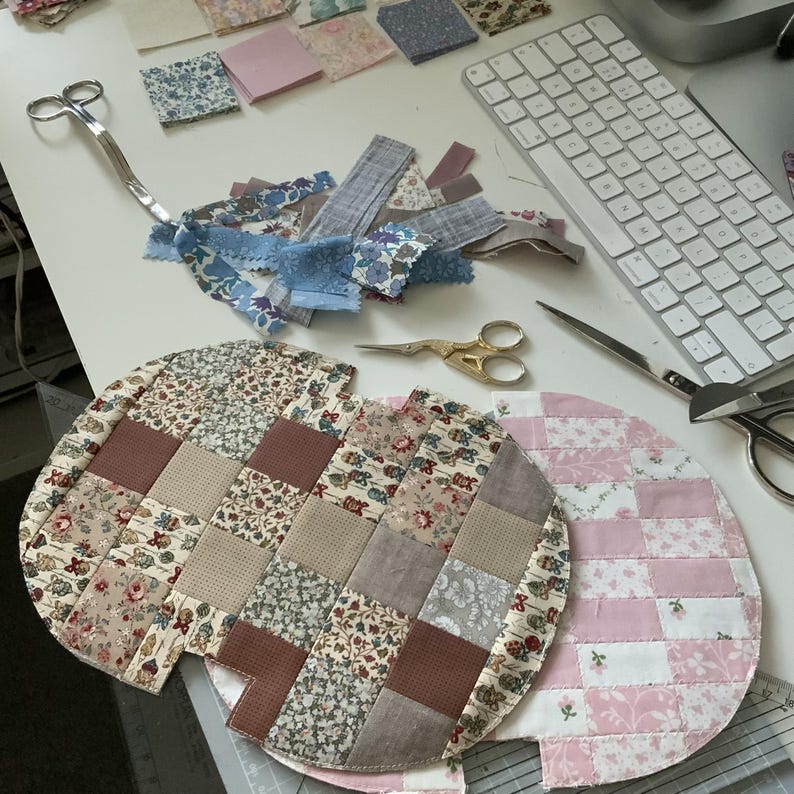 May include: A craft workspace with fabric scraps, scissors, and two quilted, circular placemats. The placemats feature patchwork designs in floral and solid fabrics. Various fabric pieces and scissors are scattered on the white surface.