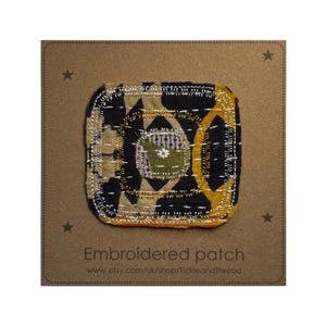 May include: A square, embroidered patch featuring a geometric pattern in black, gold, and tan, stitched with white thread. The patch has a small green central element. The text "Embroidered patch" is on the brown card.