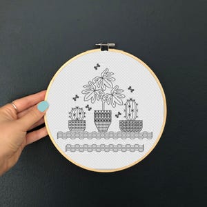 May include: A black and white cross-stitch embroidery hoop showing three potted plants: two cacti and a leafy plant. The design is set against a white background with a wavy border and small decorative elements. The hoop is held by a hand.