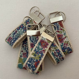 May include: A set of five keychains with floral patterned fabric and silver key rings. The fabric is a light beige with a floral pattern in shades of blue, pink, and green.