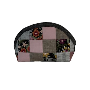 May include: A patchwork cosmetic bag with a semi-circular shape. The bag features a variety of fabric squares in pink, grey, and floral patterns. The top edge is trimmed with a dark grey zigzag stitch.