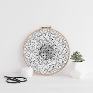 May include: A black and white floral embroidery hoop with a sunflower design. The embroidery is set in a wooden hoop. A small succulent in a white geometric pot, a ball of white thread, and black scissors are also present.