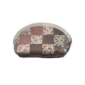 May include: An oval cosmetic bag with a patchwork design. The bag is made from squares of floral and solid fabrics in pink, brown, and cream. It has a white zip and trim, and is designed for storing makeup.