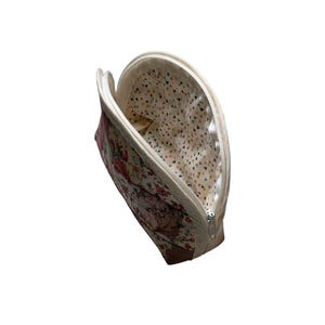 May include: A floral print cosmetic bag with a cream-coloured zip and trim. The bag's exterior features a pattern of pink and red flowers on a cream background. The interior lining has a geometric triangle pattern.