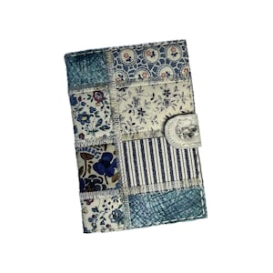 May include: A patchwork fabric wallet with a white snap closure. The wallet features various blue and white floral and striped patterns. The design includes a mix of floral prints, stripes, and textured fabrics, creating a unique, handcrafted look.