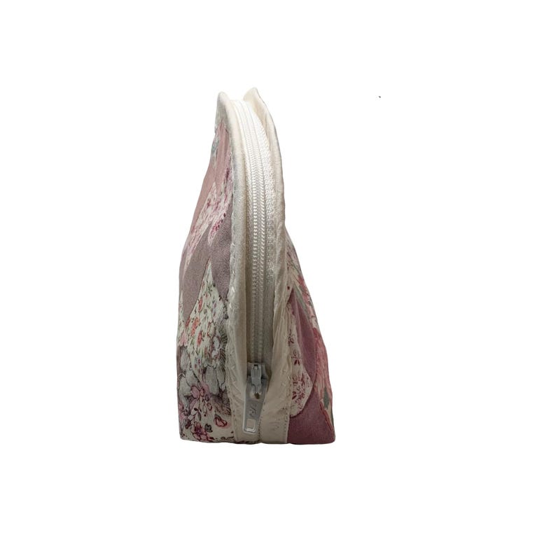 May include: A quilted cosmetic bag with a triangular shape. The bag features a patchwork design with floral and geometric patterns in shades of pink, white, and grey. A white zip runs along the top edge.