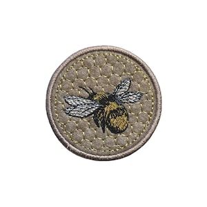 May include: Embroidered patch with a bumblebee design. The bee has black and yellow stripes and silver wings. The bee is on a tan honeycomb pattern, with a stitched border. A decorative item for clothing or accessories.