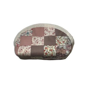 May include: An oval cosmetic bag with a patchwork design. The bag is made from squares of floral and solid fabrics in pink, brown, and cream. It has a white zip and trim, and is designed for storing makeup.