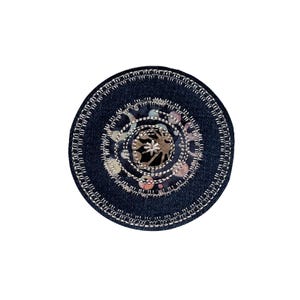 May include: A circular, dark blue denim coaster with an embroidered design. The centre features a floral pattern in muted colours, surrounded by concentric circles of stitching. The outer edge is finished with a detailed, silver-coloured stitch.