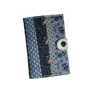 May include: A patchwork fabric wallet with a quilted design. The wallet features vertical stripes of blue floral patterns and a dark floral pattern. It has a white tab closure with a green button.