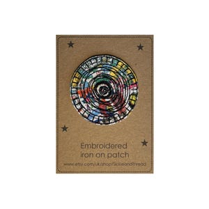 May include: A circular embroidered iron-on patch featuring a multi-coloured floral pattern overlaid with concentric black stitched circles. The patch is mounted on a rectangular brown card printed with the text Embroidered iron on patch.