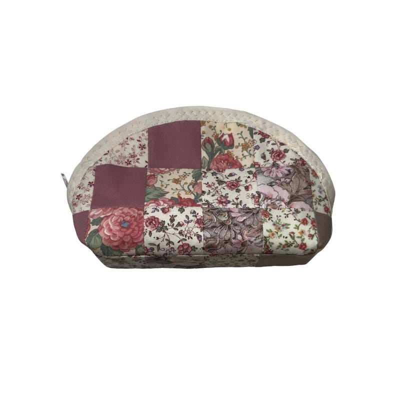 May include: A quilted cosmetic bag with a patchwork design. The bag features squares of floral fabric in shades of pink, cream, and burgundy. The top edge is trimmed with cream-coloured lace. A small zip is visible on the left side.