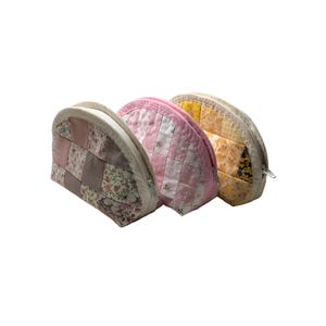 May include: Three quilted cosmetic bags. The bags are in shades of pink, yellow, and neutral tones. Each bag has a different patchwork design and a zip closure. The bags are made of fabric with floral patterns.