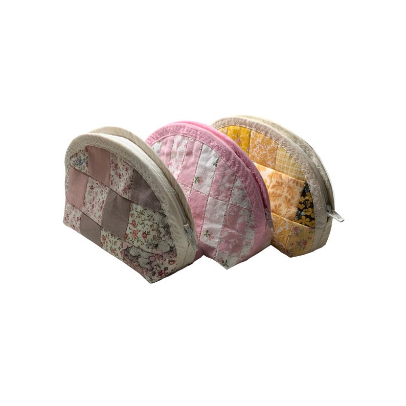 May include: Three quilted cosmetic bags. The bags are in shades of pink, yellow, and neutral tones. Each bag has a different patchwork design and a zip closure. The bags are made of fabric with floral patterns.