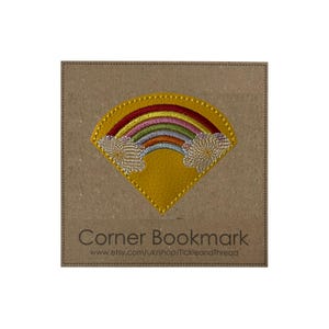 May include: A yellow corner bookmark featuring a rainbow design and embroidered clouds. The bookmark is on a brown card with the text "Corner Bookmark" and a website address. The rainbow has red, orange, yellow, green, blue, and purple arches.