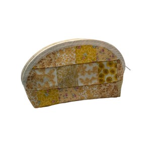 May include: A half-moon shaped cosmetic bag with a patchwork design. The bag features a variety of yellow and tan floral patterns, with a cream-coloured lace trim along the top edge. The zipper is silver.