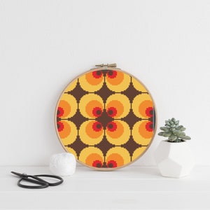 Może przedstawiać: Okrągła rama do haftu z brązowym drewnianym obramowaniem i kolorowym geometrycznym wzorem. Wzór zawiera powtarzający się wzór pomarańczowych, żółtych i czerwonych kół z czarnymi środkami.