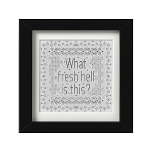 Op de afbeelding: Een ingelijst kruissteekkunstwerk met de tekst "What fresh hell is this?" in zwart garen. De tekst staat centraal op een witte achtergrond, omgeven door een decoratieve rand. De lijst is zwart.