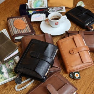 May include: An assortment of leather journals and notebooks in various colors, including black, brown, and tan. A small cup of espresso sits on a white saucer. The journals feature clasps, straps, and decorative details.