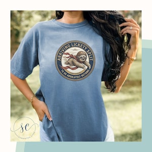 Könnte beinhalten: Ein blaues T-Shirt mit kurzen Ärmeln und einem kreisförmigen Aufdruck eines Faultiers mit Fliegerbrille und rotem Schal. Der Text lautet ARRIVING LICKETY SPLIT TO DO ABSOLUTELY NOTHING. Das Design enthält die Silhouette eines kleinen Flugzeugs.