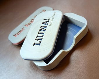 Puede incluir: Una caja blanca con un interior azul y una tapa que dice "LIUNA! Feel the Power" en una fuente estilizada. La caja está hecha de un material texturizado y tiene una forma redondeada.