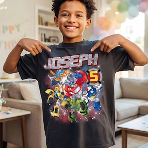 May include: A dark gray t-shirt featuring the name "JOSEPH" in a silver, textured font above a colorful graphic of cartoon robots. The number "5" is in a circle on the right side of the graphic. The boy is pointing at the shirt.