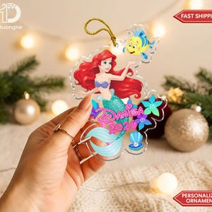 May include: A clear acrylic ornament featuring a colorful illustration of a mermaid with long red hair, a yellow fish, and blue flowers. The name Danisa and the year 2026 are printed in pink script. A gold string is attached to the top for hanging.