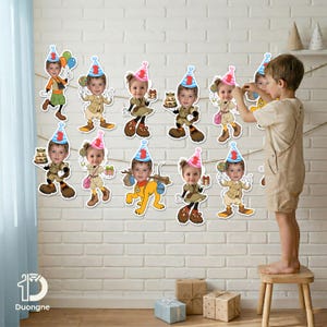 May include: A garland of birthday party decorations featuring cartoon images of a child with party hats, holding gifts, balloons, and candy. The decorations are strung across a white brick wall. A child is standing on a small wooden stool.