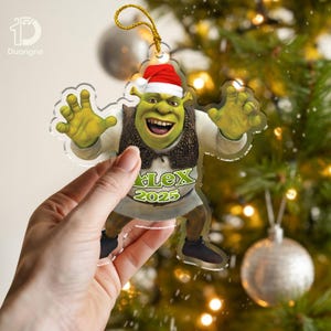 Pode incluir: Um enfeite de Natal de acrílico transparente com Shrek usando um gorro de Papai Noel. O enfeite tem o texto "Lex 2025" em verde. O enfeite está pendurado em uma corda dourada, com uma árvore de Natal ao fundo.