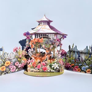 May include: A 3D paper art scene depicting a gazebo, deer, and colourful flowers. The gazebo has a pink roof and is surrounded by vibrant blooms. Two deer stand inside the gazebo. White picket fences frame the floral garden, creating a whimsical, nature-themed display.
