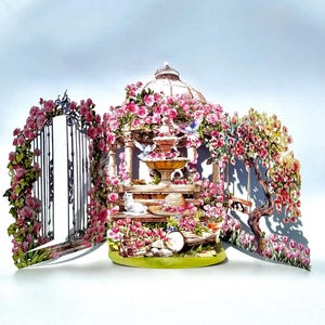 May include: A detailed paper diorama of a garden scene with a gazebo, fountain, and gate, all adorned with pink roses. A white cat sits on a bench near a straw hat. The scene is surrounded by flowering trees and tulips.