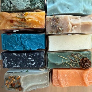 May include: A collection of handmade soaps in various colors and textures. The soaps are arranged in rows, showcasing a variety of hues including green, blue, orange, and beige. Some soaps are decorated with dried herbs and a small pine cone.