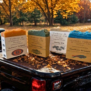May include: Four bars of handmade soap in various colors, including orange, green, beige, and blue, each with a unique label. The soaps are displayed on the back of a truck, with an autumn-themed background.