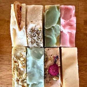May include: Eight rectangular bars of handcrafted soap arranged in two rows. The bars feature various textures and inclusions, including a dried orange slice, lavender buds, dried flower petals, and smooth surfaces in shades of white, pink, green, and blue.