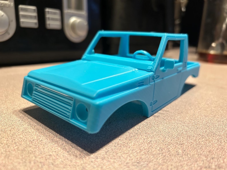 May include: A light blue plastic model car chassis, a bare frame of a pickup truck with no body panels, wheels, or engine. The model is missing the doors and hood, but the windshield frame is intact. The model is sitting on a light gray surface.