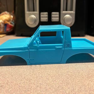 May include: A light blue plastic model car body, a pickup truck with a single cab and a flatbed. The model has a door on the driver's side and a 3L printed on the bed.