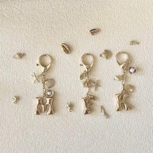 Op de afbeelding: Goudkleurige sleutelhangers met de letters M, E en K. Elke sleutelhanger heeft een karabijnhaak, bedels van schelpen, zeesterren en parels. Extra schelp- en zeesterbedels zijn verspreid over een crèmekleurig oppervlak.