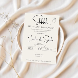 Puede incluir: Una tarjeta de invitación a una fiesta de compromiso con el texto "Shhh... He's Going to Propose!" en escritura negra. La tarjeta también incluye los nombres "Carlie & Jake" y los detalles del evento. La tarjeta está sobre un plato blanco.