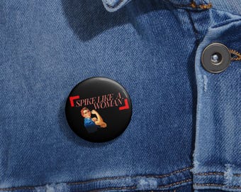 May include: A black button with a red border and the text "Spike Like A Woman" in white. The button features a vintage-style illustration of a woman flexing her bicep.
