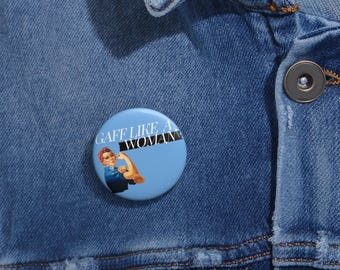 May include: A blue button with a white background and the text "GAFF LIKE A WOMAN" in black. The button features a woman in a blue dress with a red bandana, flexing her bicep.