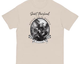 May include: A beige t-shirt featuring a black and white graphic of Saint Michael, with the text "Saint Michael, Defend Us in Battle, The Archangel". The design includes an oval frame with roses.