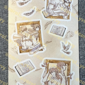 May include: A sticker sheet featuring anime-style illustrations of characters from Attack on Titan, including Erwin Smith, Levi Ackerman, Hans Zoe, Eren Yeager, and Mikasa Ackerman, framed in gold, alongside drawings of open books, feathers, and birds.