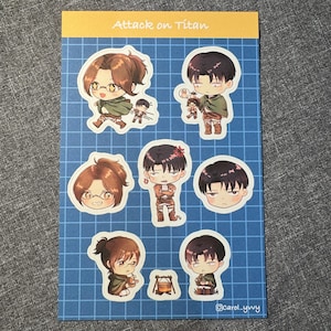 May include: A rectangular sticker sheet with a blue grid background and the text Attack on Titan at the top. It features seven individual chibi-style character stickers depicting figures with brown and black hair, wearing green capes and brown gear.